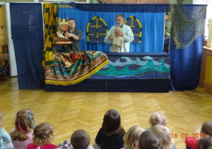 Scenografia teatralna - morze, oraz zamek. Dwóch aktorów z dwomalalkami - królową i rybakiem. Dzieci oglądające przedstawienie.